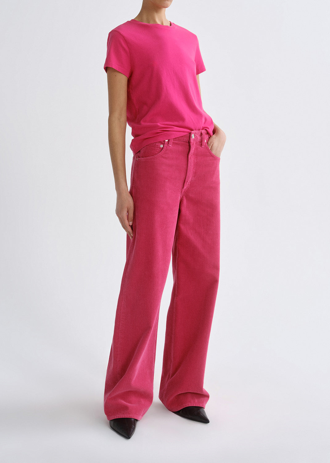 Person wearing a bright pink short sleeve crewneck tee with bright pink straight leg jeans viewed from the side on a grey background