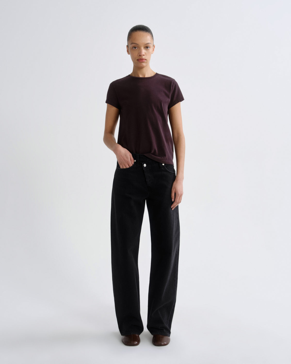 A model stands against a light gray studio background wearing a dark plum short-sleeve t-shirt and black wide-leg trousers.
