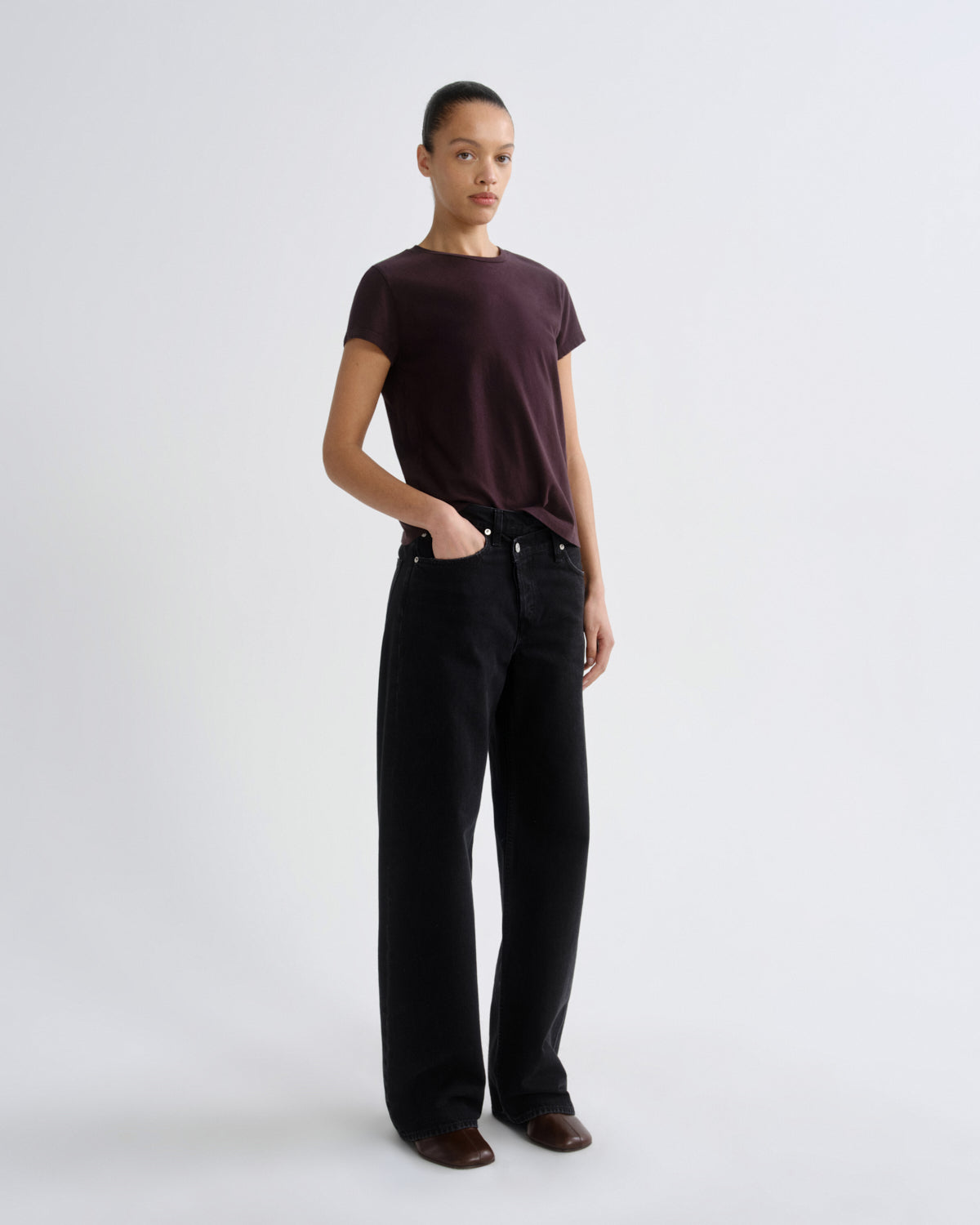 A model stands against a light gray studio background wearing a dark plum short-sleeve t-shirt and black wide-leg jeans.