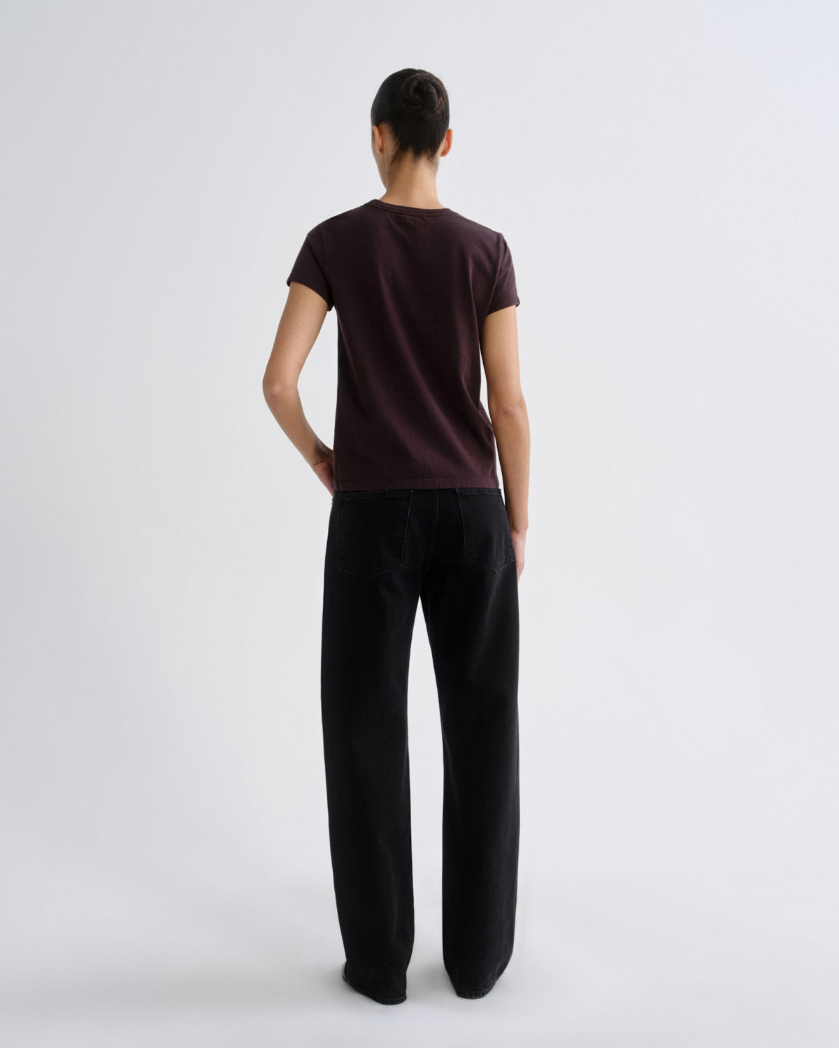 A model viewed from the back wearing a dark plum short-sleeve t-shirt and black wide-leg trousers, standing against a light gray studio background.