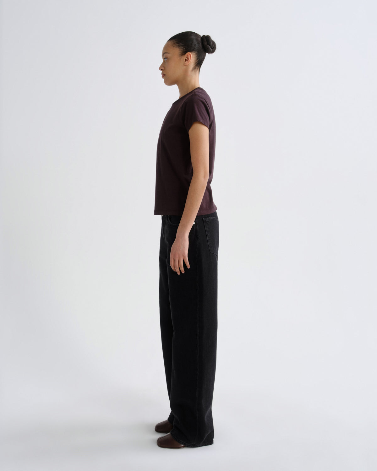 A model standing in profile wearing a dark plum short-sleeve t-shirt and black wide-leg trousers against a plain light gray studio background.