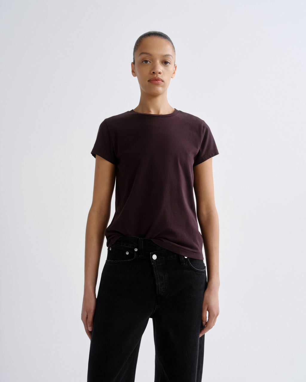 A model wearing a dark plum short-sleeve crewneck t-shirt tucked into black jeans, standing against a light gray studio background.