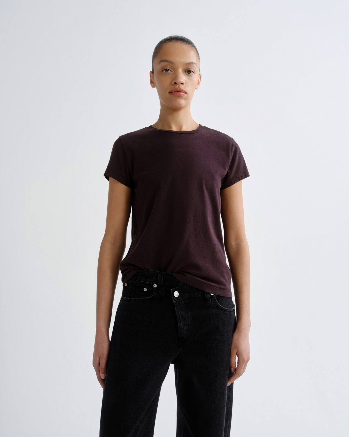 A model wearing a dark plum short-sleeve crewneck t-shirt tucked into black jeans, standing against a light gray studio background.