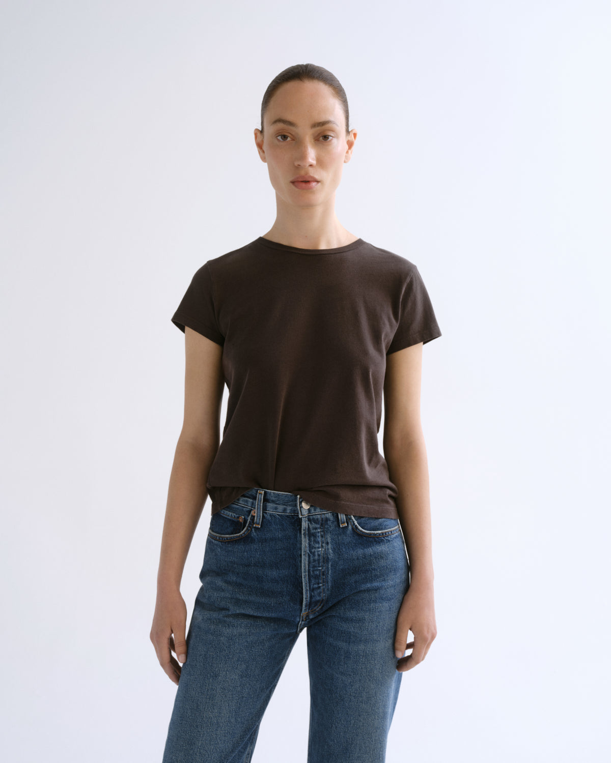 A woman modeling a dark brown short-sleeve crewneck t-shirt paired with blue jeans, standing against a plain light gray studio background.