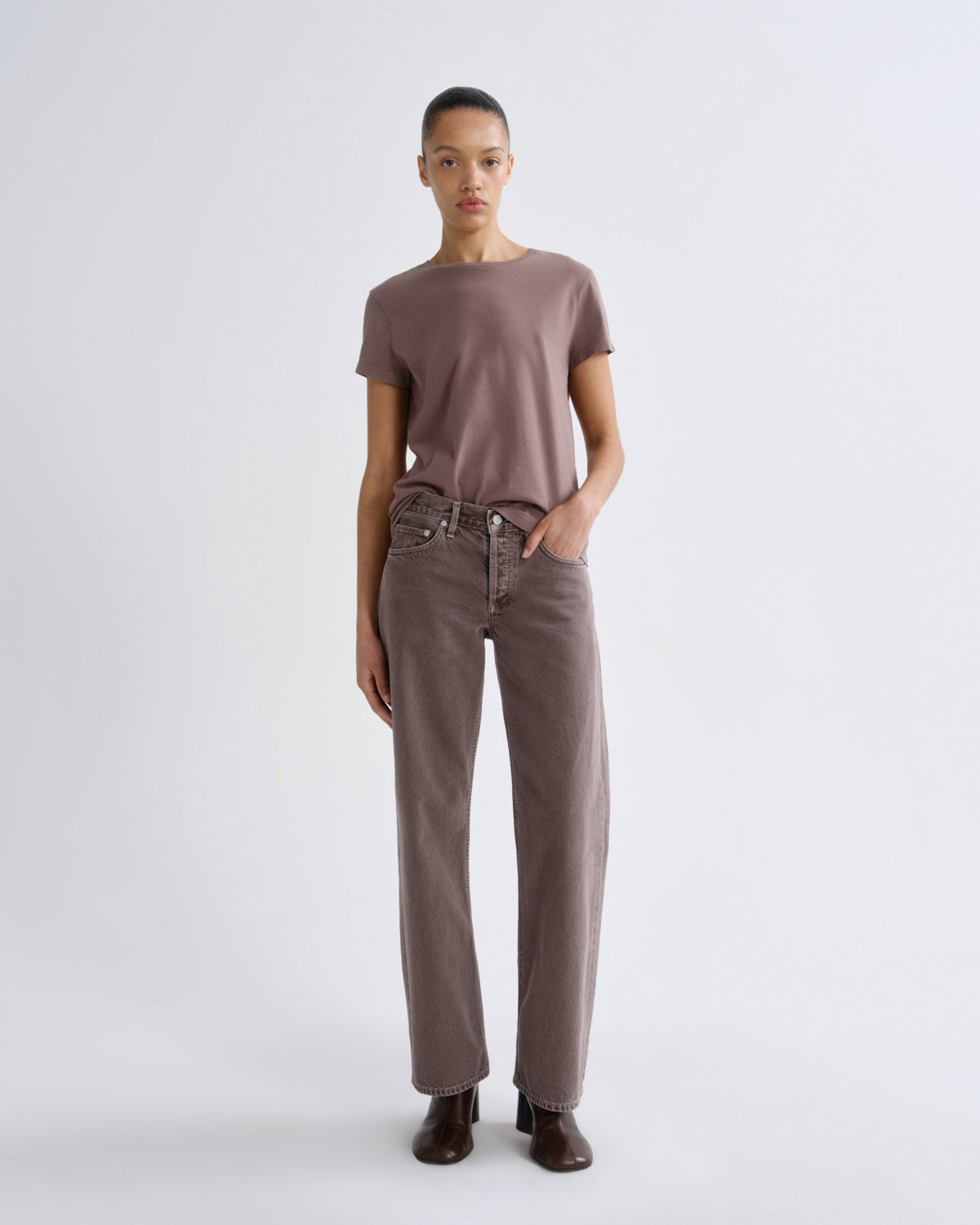 A model stands against a light gray studio background wearing a muted mauve-brown short-sleeve crewneck top, matching wide-leg jeans, and dark brown heeled shoes.