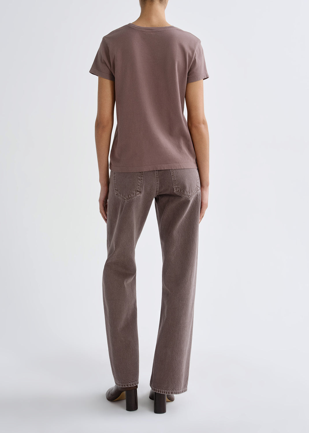 Person wearing a mauve short sleeve crewneck tee with mauve straight leg jeans viewed from the back on a grey background