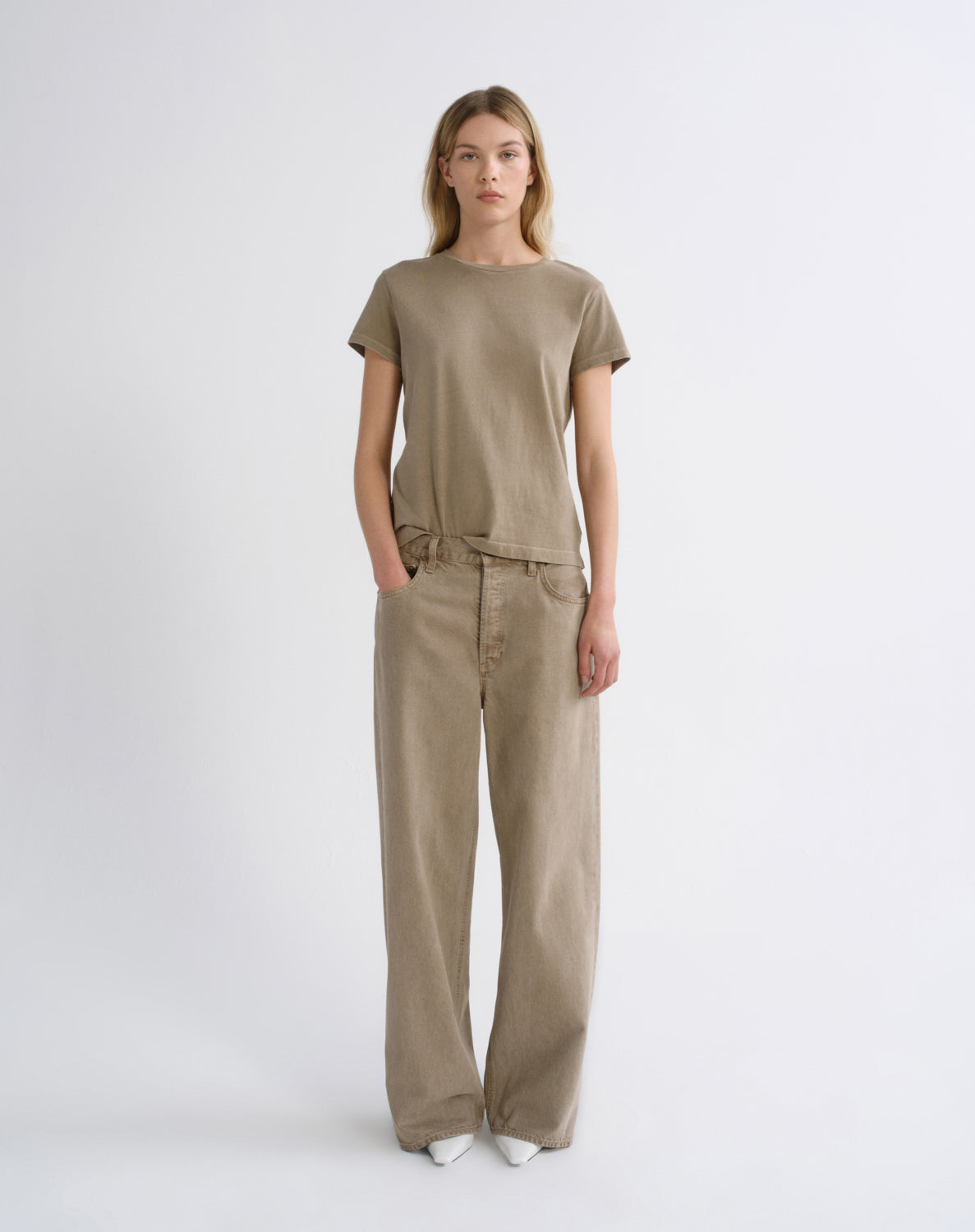 A woman stands against a plain light gray studio background wearing a loose short-sleeve t-shirt and matching wide-leg pants in a warm taupe-beige color.