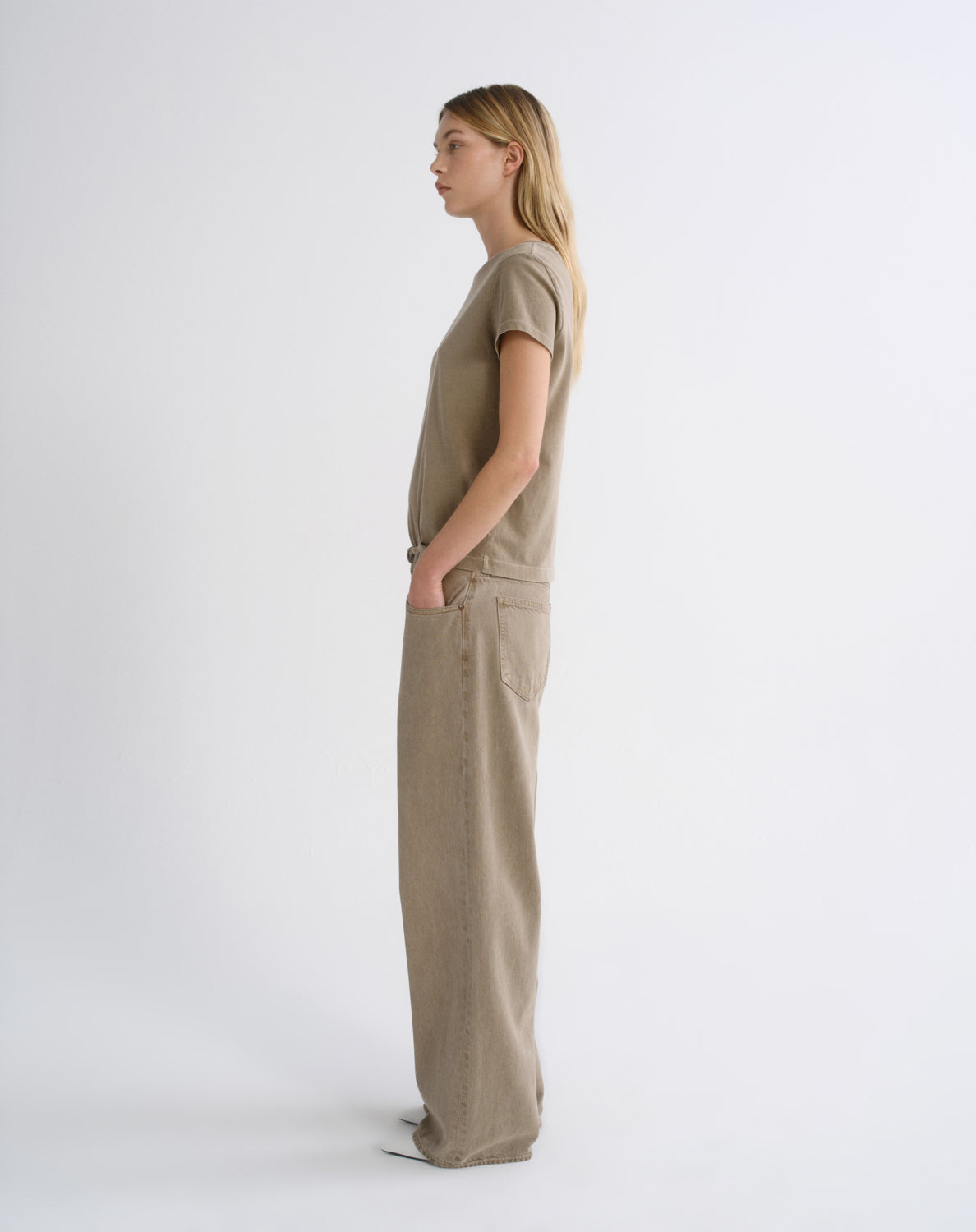 A woman stands in profile wearing a matching taupe knit short-sleeve top and wide-leg trousers set, photographed against a clean white studio background.