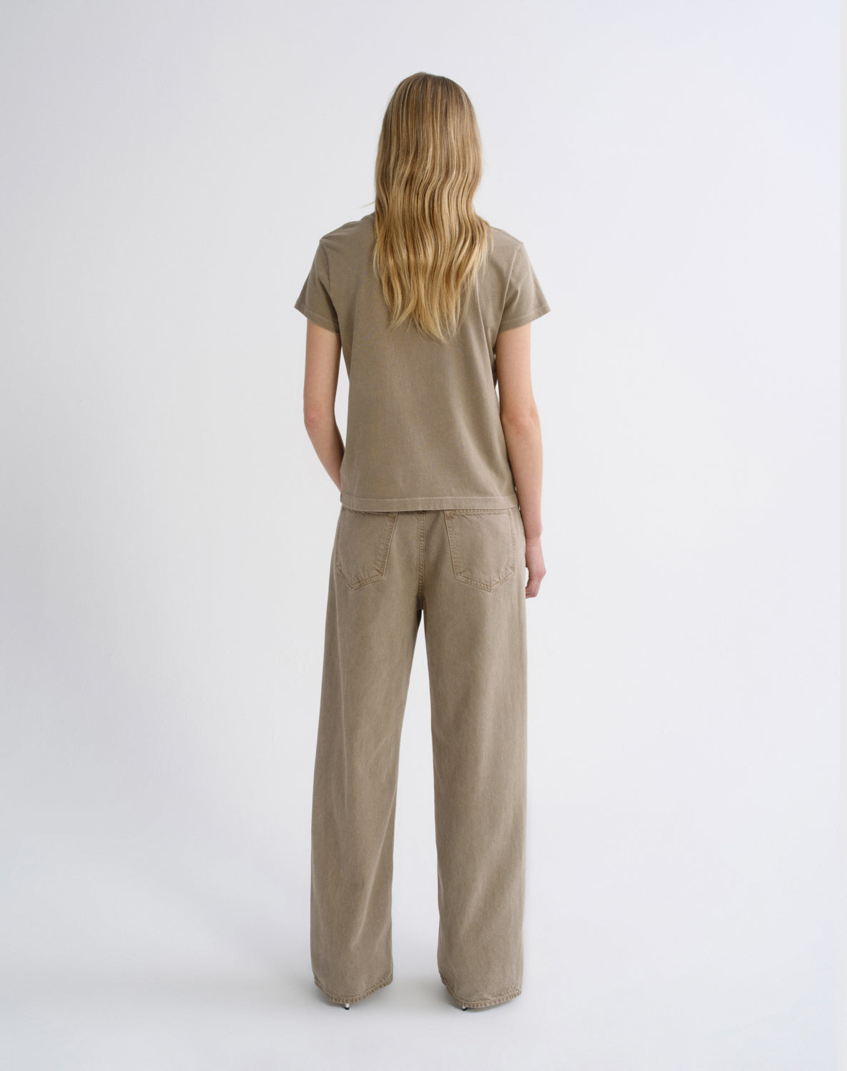 A woman viewed from behind wearing a matching beige short-sleeve t-shirt and wide-leg trousers set against a plain light gray studio background.