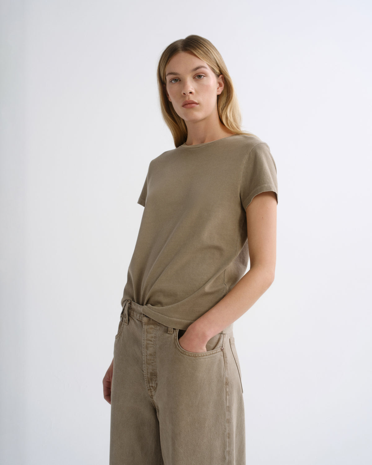 A woman modeling a short-sleeve t-shirt and matching wide-leg trousers in a solid taupe-beige color, standing against a plain light gray studio background.