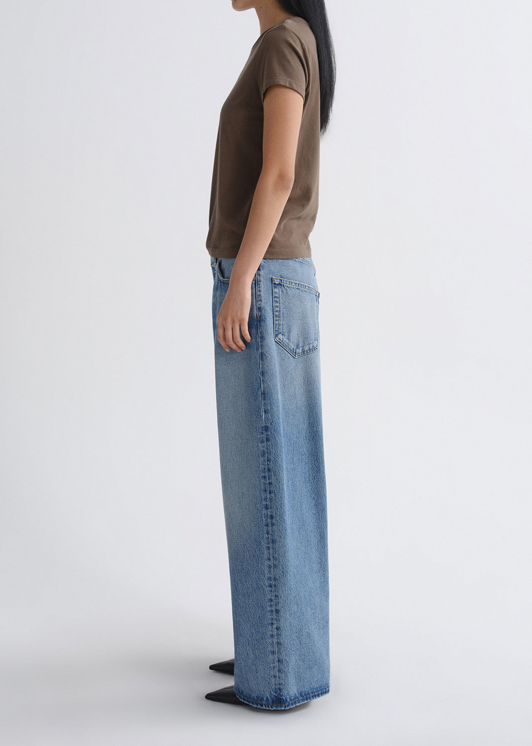 Person wearing a brown short sleeve crewneck tee with light wash wide leg jeans viewed from the side on a grey background