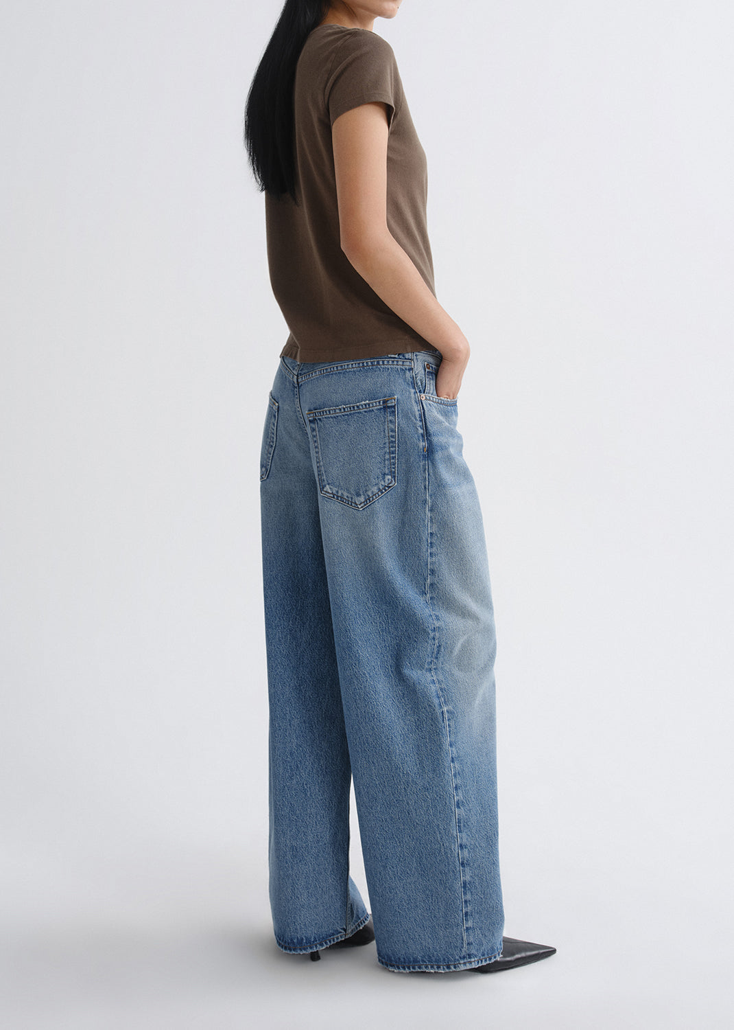 Person wearing a brown short sleeve crewneck tee with light wash wide leg jeans viewed from the side on a grey background