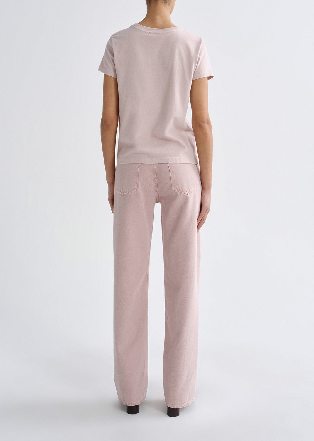 Person wearing a light pink short sleeve crewneck tee with light pink straight leg jeans viewed from the back on a grey background