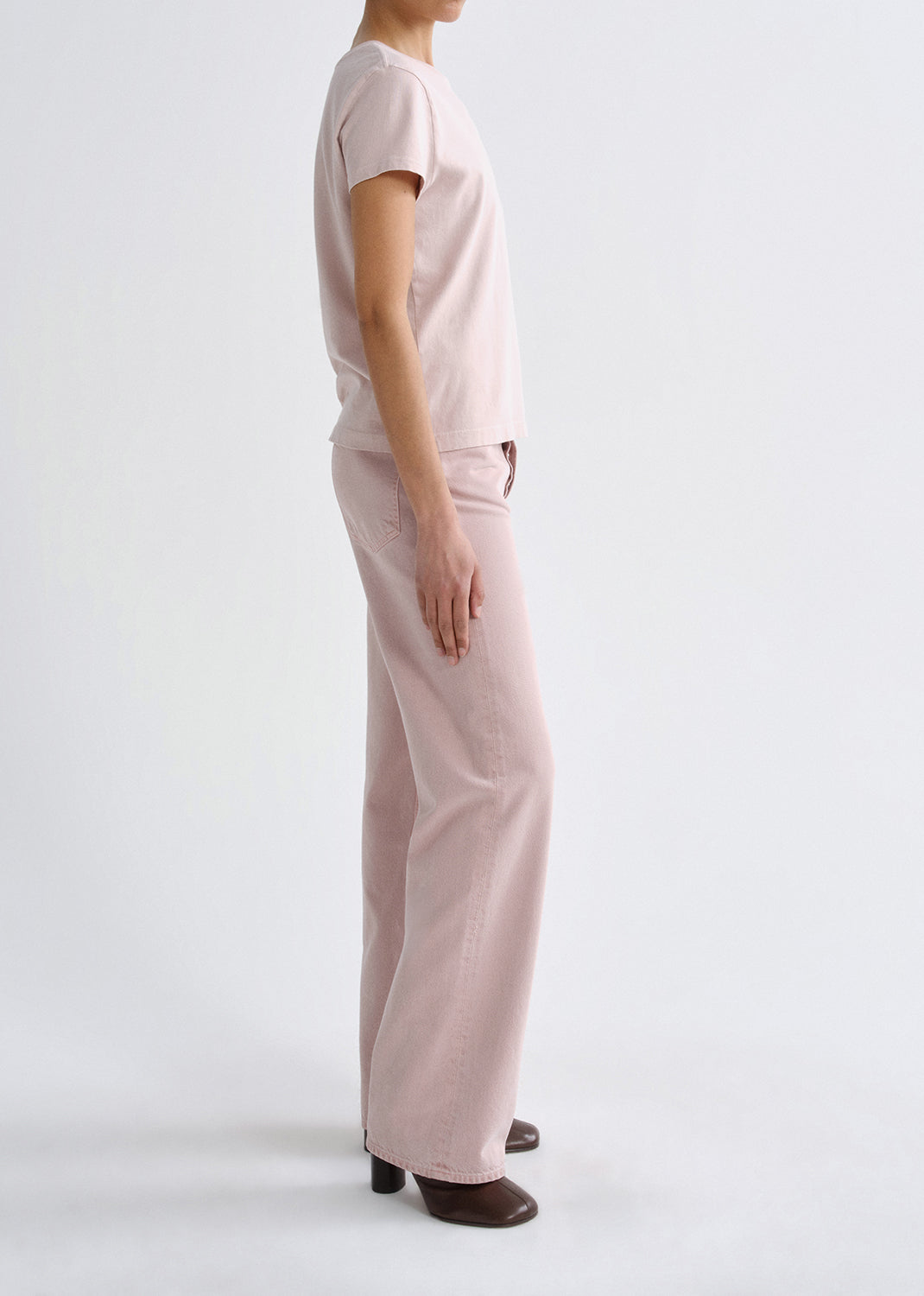 Person wearing a light pink short sleeve crewneck tee with light pink straight leg jeans viewed from the side on a grey background