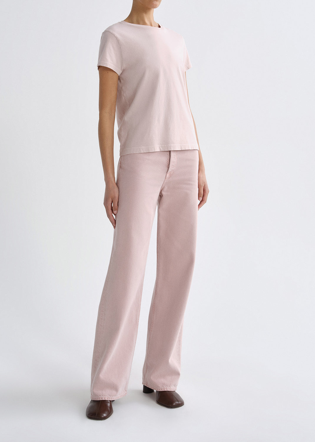 Person wearing a light pink short sleeve crewneck tee with light pink straight leg jeans viewed from the side on a grey background