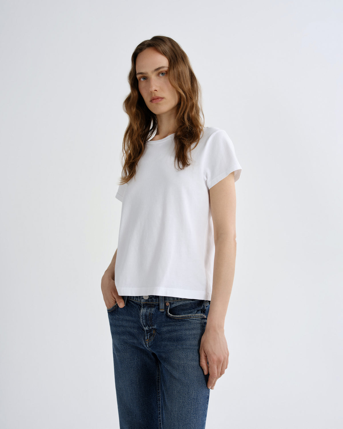 A woman modeling a white short-sleeve crewneck t-shirt paired with dark blue jeans, standing against a plain light gray studio background.