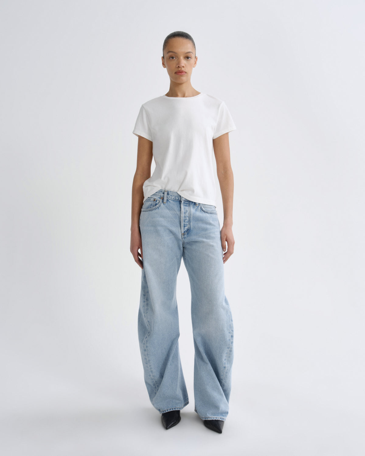 A model stands against a light gray studio background wearing a white short-sleeve crewneck t-shirt and light-wash wide-leg jeans.