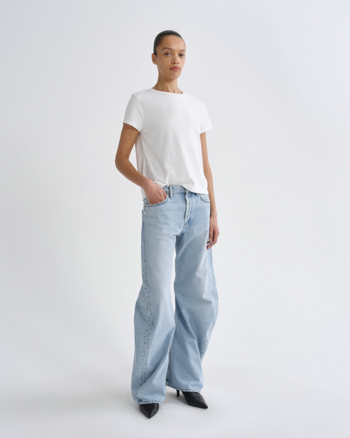 A model stands against a light gray studio background wearing a white short-sleeve crewneck t-shirt, light-wash wide-leg jeans, and black pointed-toe heels.