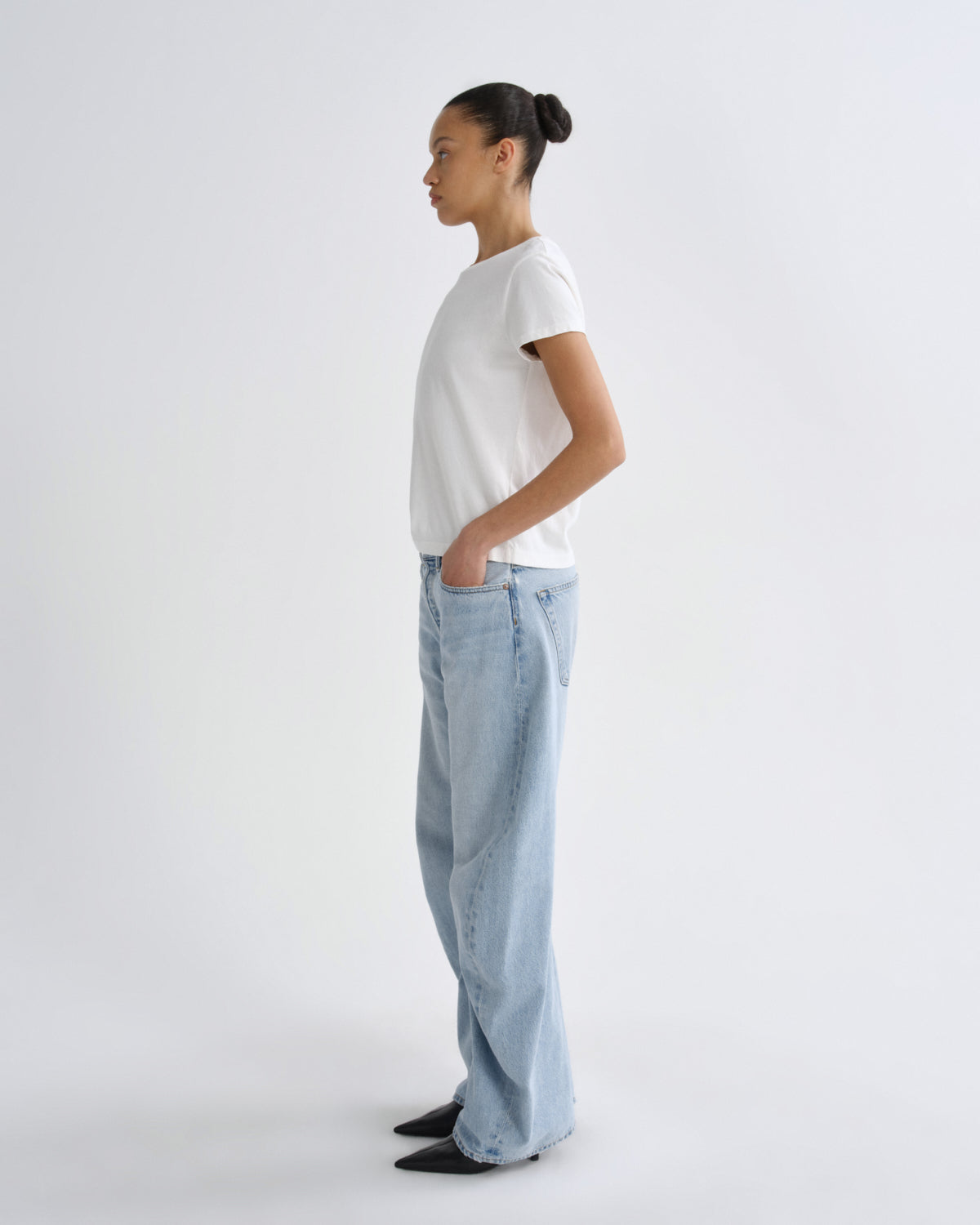 A model standing in profile wearing a white short-sleeve t-shirt, light-wash wide-leg jeans, and black pointed shoes against a plain light gray studio background.