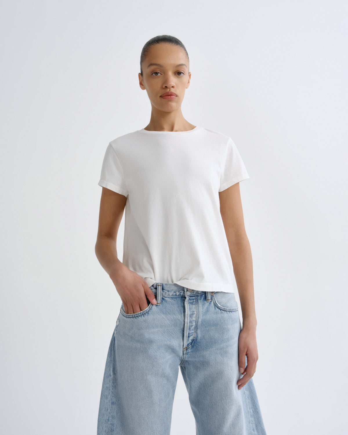 A model wearing a white short-sleeve crewneck t-shirt and light-wash blue jeans, standing against a plain light gray studio background.