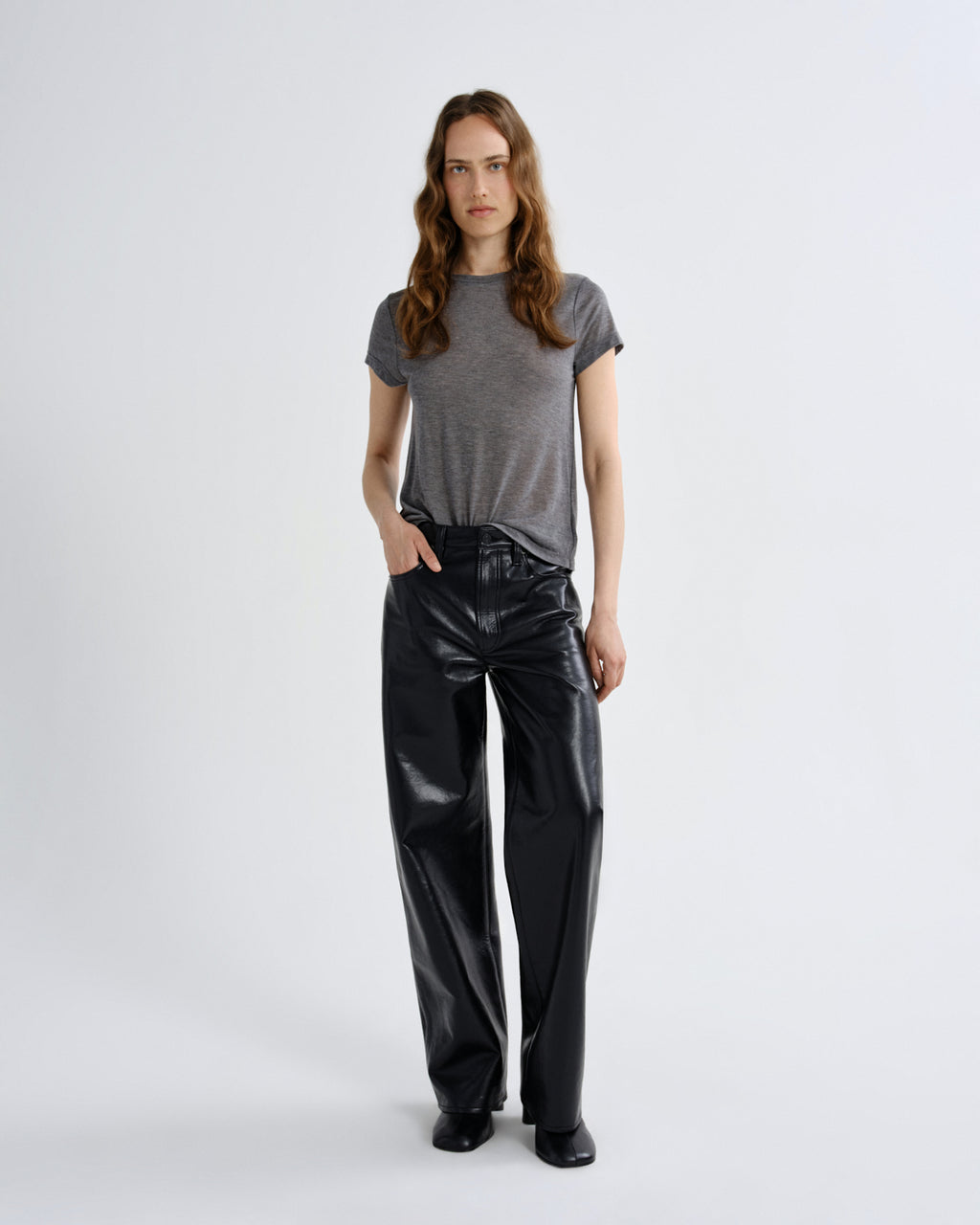 A woman stands against a light gray studio background wearing a short-sleeve gray t-shirt and shiny black wide-leg pants.