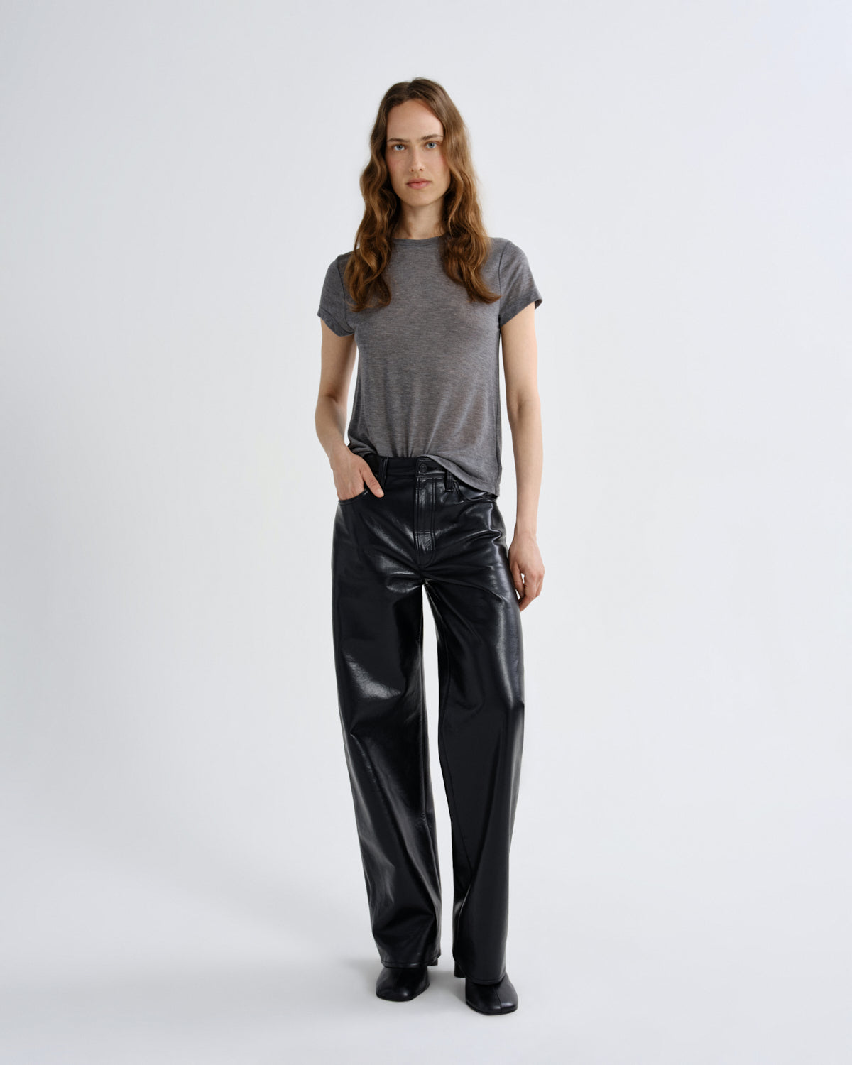A woman stands against a light gray studio background wearing a short-sleeve gray t-shirt and shiny black wide-leg pants.