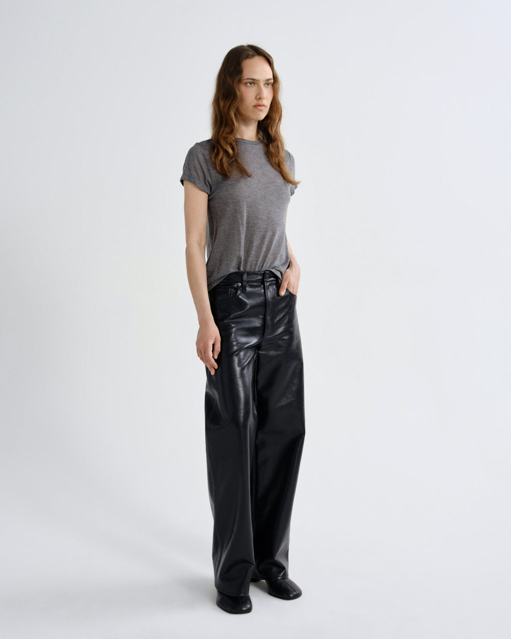 A woman stands against a light gray studio background wearing a short-sleeve gray t-shirt and shiny black wide-leg pants.