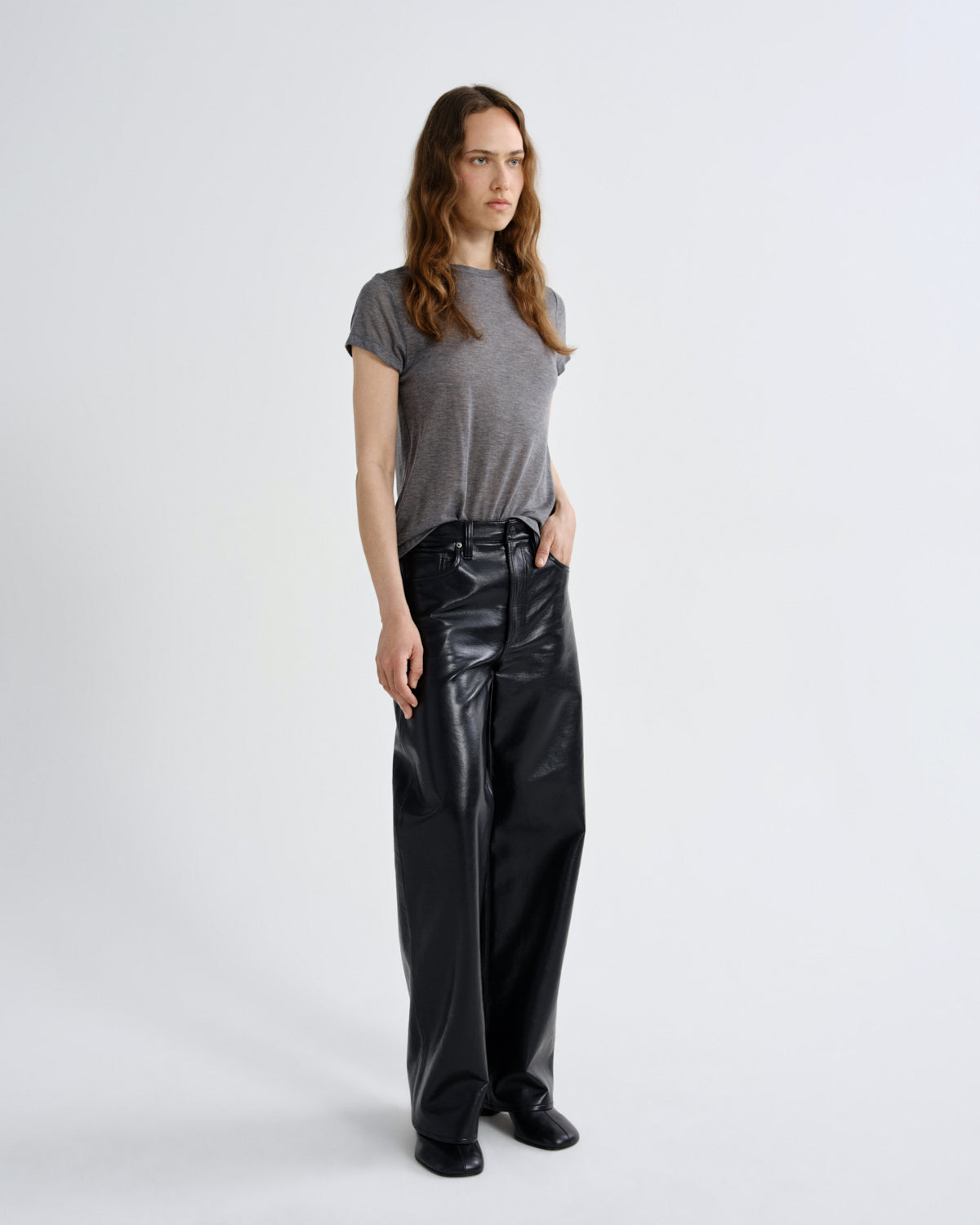 A woman stands against a light gray studio background wearing a short-sleeve gray t-shirt and shiny black wide-leg pants.