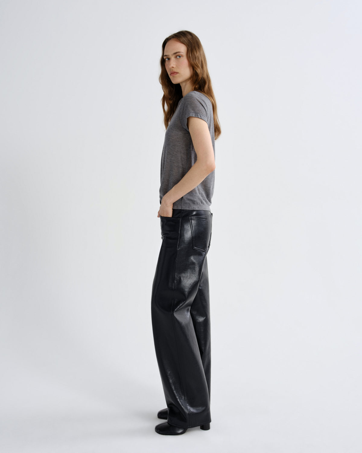 A woman stands in profile wearing a gray short-sleeve t-shirt and shiny black wide-leg pants against a plain light gray studio background.