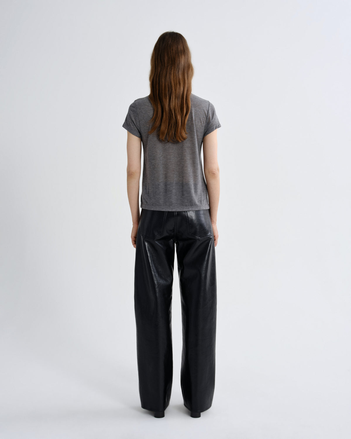 A model viewed from behind wearing a gray short-sleeve t-shirt and black wide-leg faux-leather pants, standing against a plain light gray studio background.