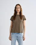 A woman modeling a short-sleeve crewneck t-shirt in a muted brownish-gray color, paired with light-wash blue jeans, against a plain light gray studio background.