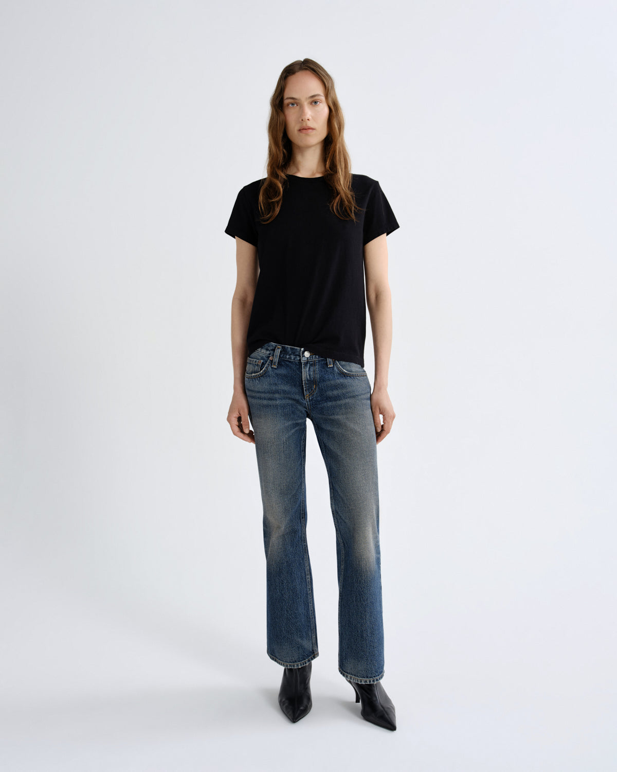 A woman stands against a light gray studio background wearing a black short-sleeve crewneck t-shirt, blue straight-leg jeans, and black pointed ankle boots.