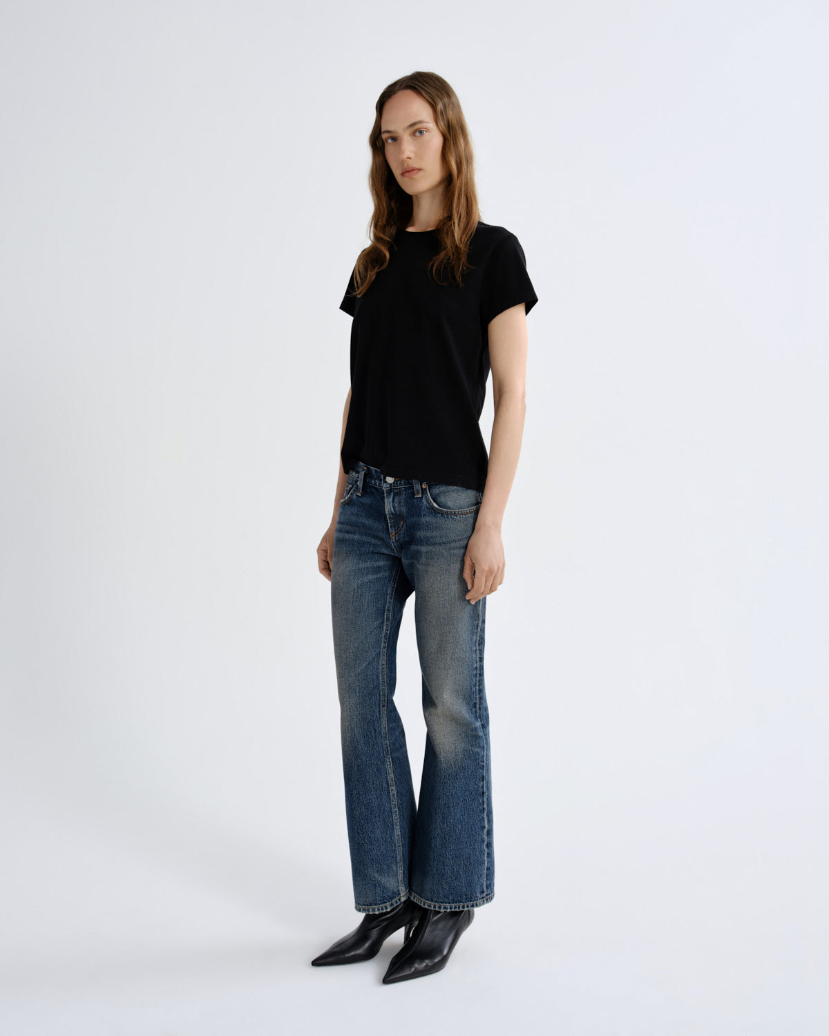 A woman modeling a black short-sleeve crewneck t-shirt and blue flare jeans with black pointed-toe ankle boots, standing against a plain light gray studio background.