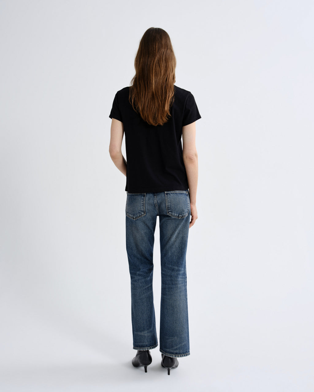 A woman viewed from behind wearing a black short-sleeve t-shirt, blue straight-leg jeans, and black pointed-toe heels against a plain light gray studio background.