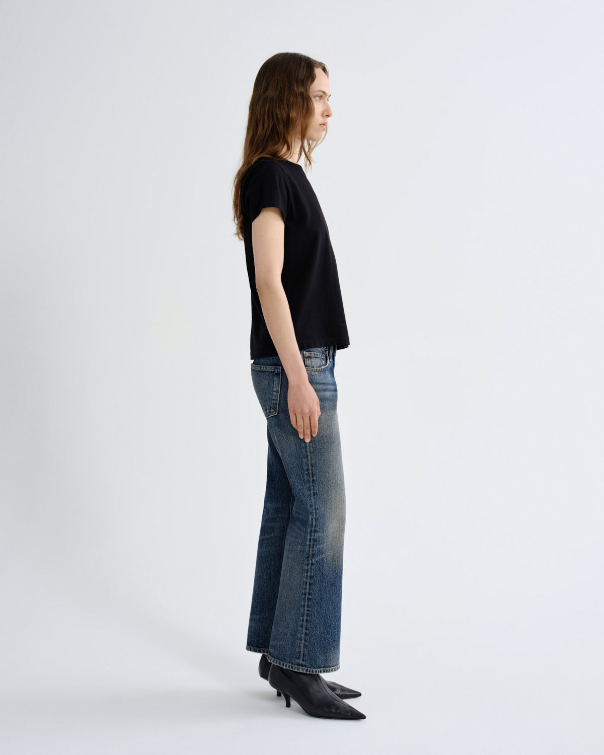 A woman stands in profile wearing a black short-sleeve top, blue wide-leg jeans, and black pointed-toe heeled boots against a plain light gray studio background.