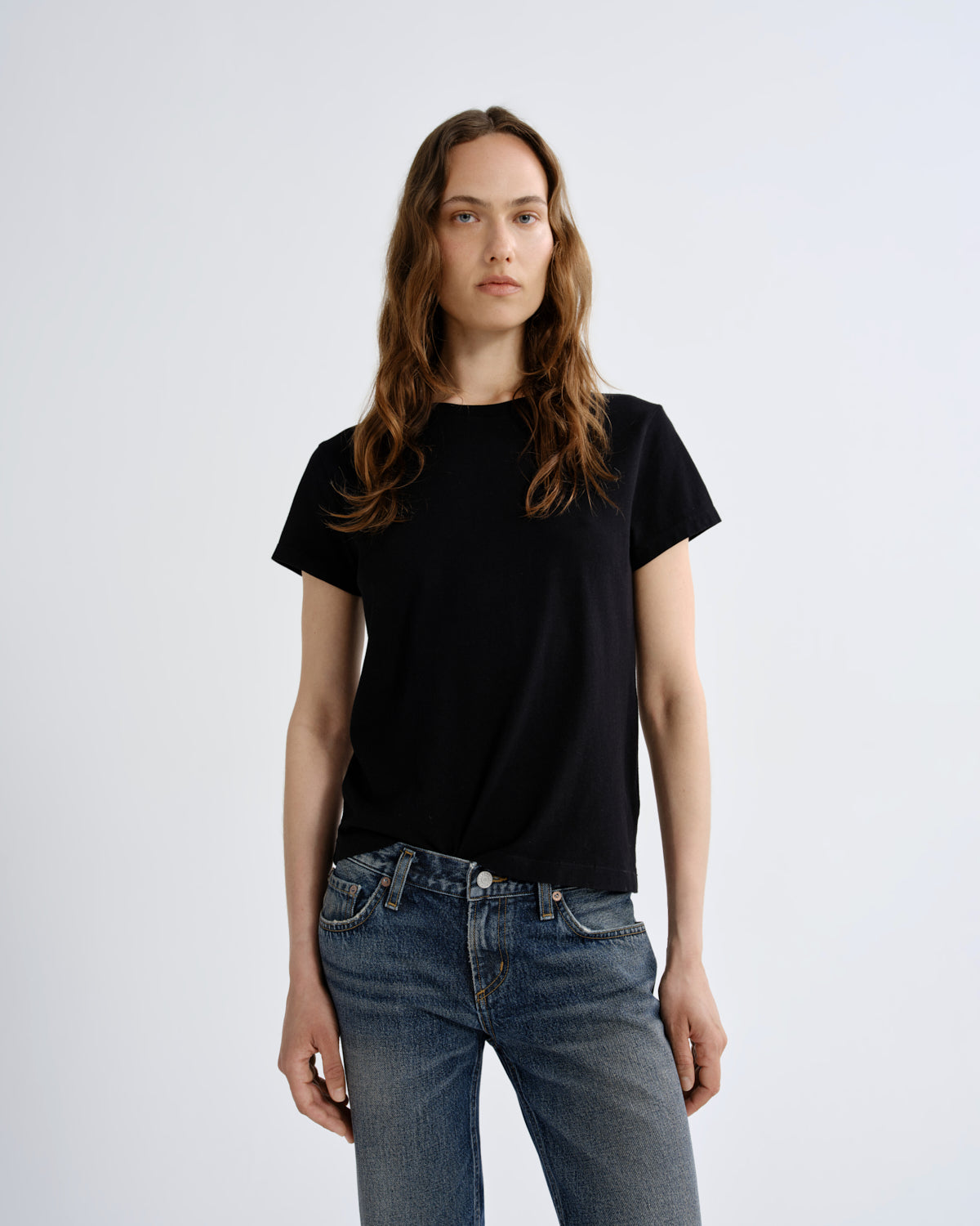 A woman modeling a black short-sleeve crewneck t-shirt paired with blue jeans, standing against a light gray studio background.