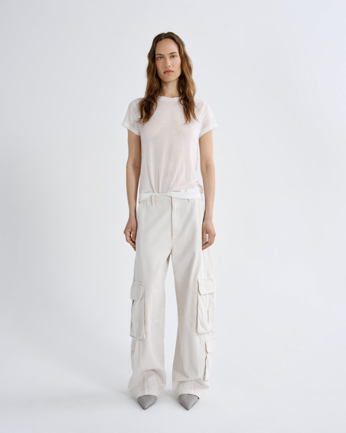 A woman stands against a light gray studio background wearing a loose white short-sleeve T-shirt and matching white wide-leg cargo pants with gray pointed shoes.