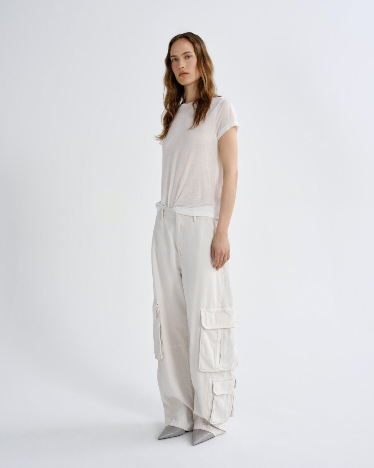 A woman modeling a light beige short-sleeve t-shirt and matching wide-leg cargo pants with silver pointed shoes, photographed against a seamless white studio background.