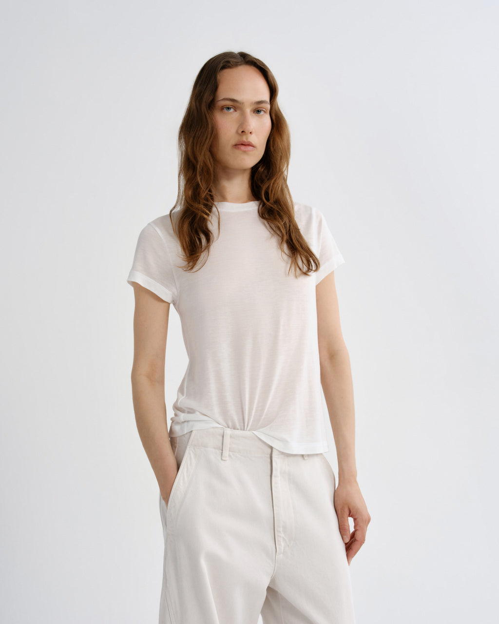 A woman modeling a short-sleeve crewneck t-shirt in off-white, tucked into matching off-white trousers, against a plain light gray studio background.