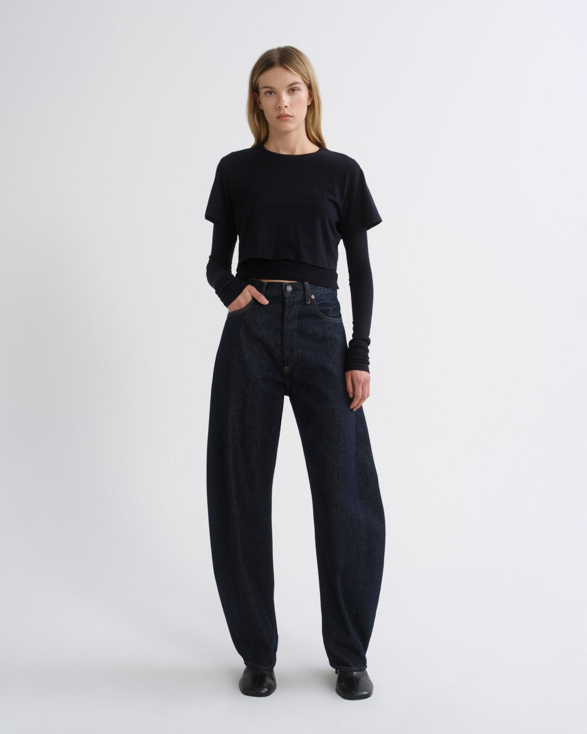 A model stands against a light gray studio background wearing a black short-sleeve cropped top over a black long-sleeve shirt, paired with dark wide-leg jeans and black shoes.