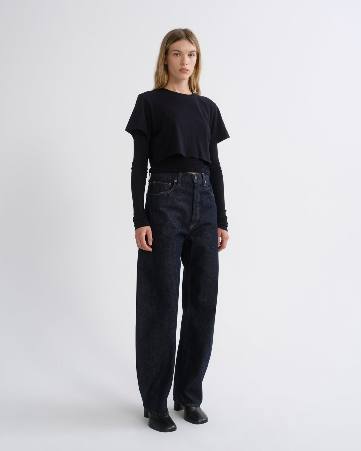 A model stands against a light gray studio background wearing a black cropped short-sleeve top over a black long-sleeve shirt, paired with dark blue wide-leg jeans and black shoes.