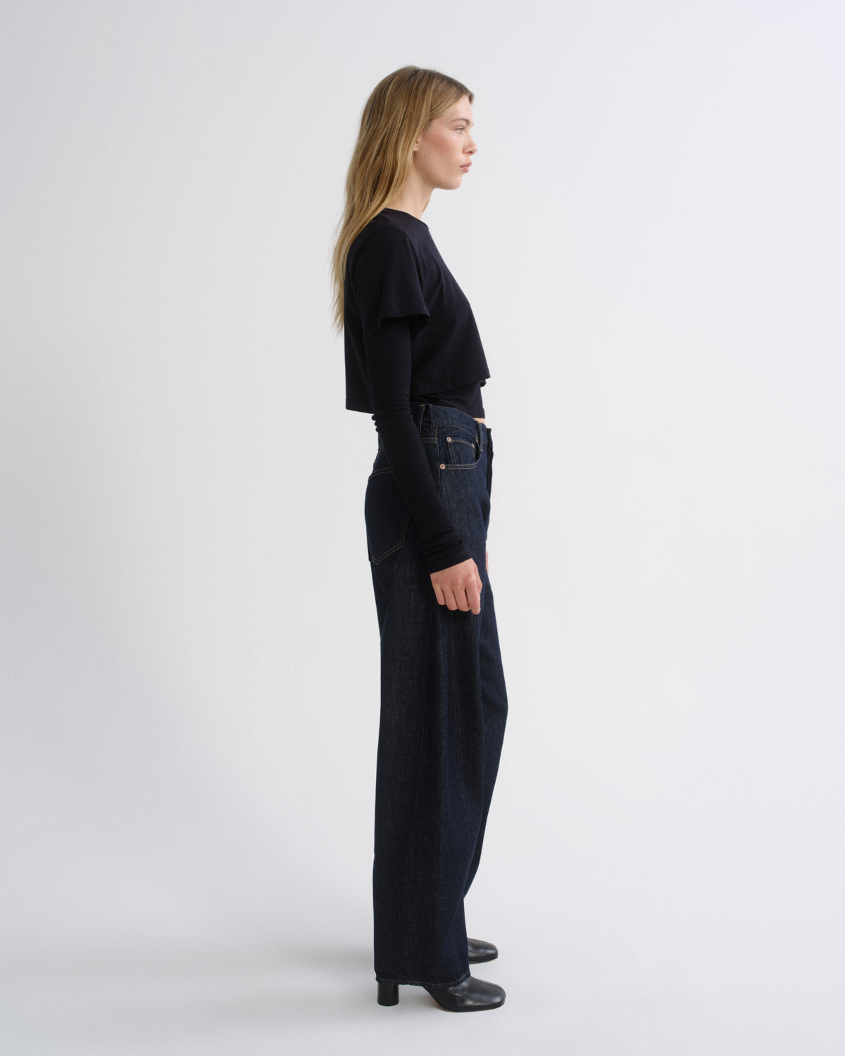 A woman stands in profile wearing a black long-sleeve top with ruffled shoulders and dark wide-leg jeans, photographed against a clean light gray studio background.