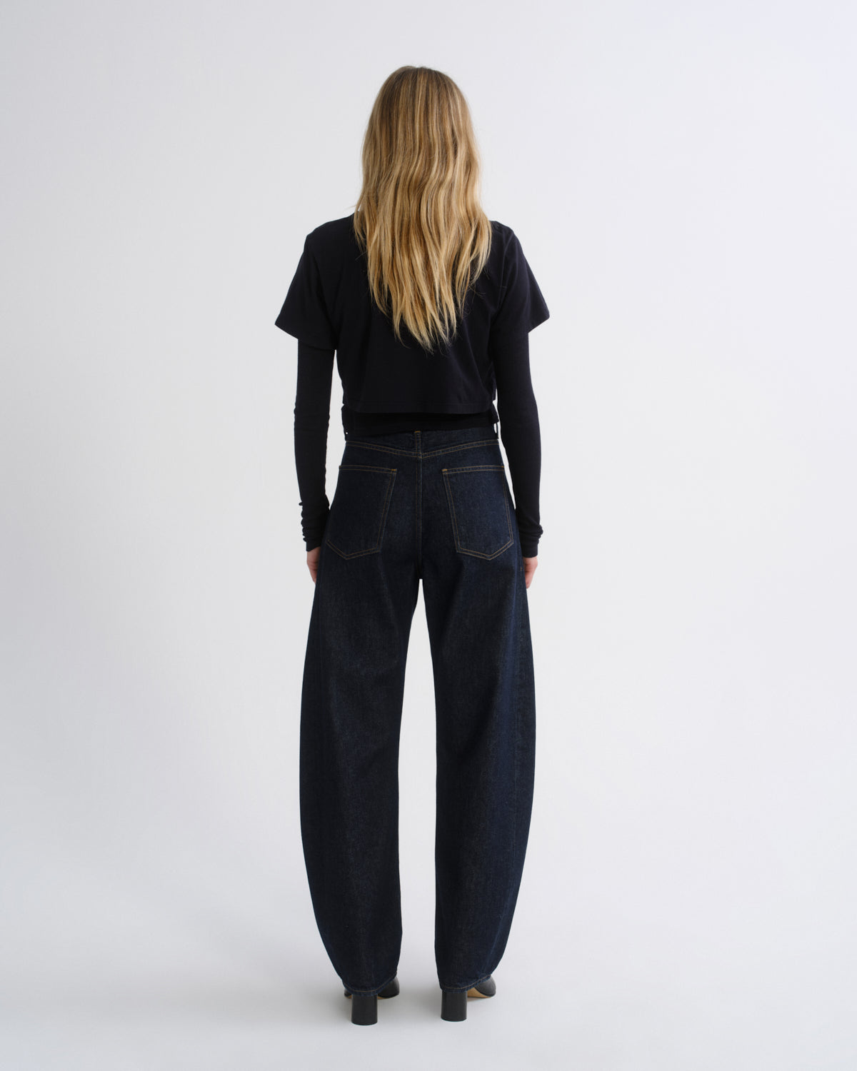 A woman viewed from behind wearing a black short-sleeve top, dark wide-leg jeans, and black shoes, standing against a plain light gray studio background.