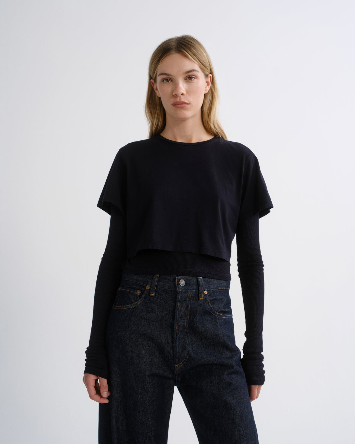 A woman modeling a black cropped long-sleeve top over a black long-sleeve undershirt, paired with dark blue jeans, against a plain light gray studio background.