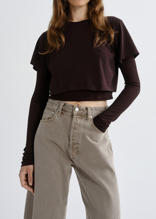 A model wearing a dark brown cropped long-sleeve t-shirt with ruffled sleeves over a matching dark brown long-sleeve base, paired with high-waisted grey wide-leg jeans.