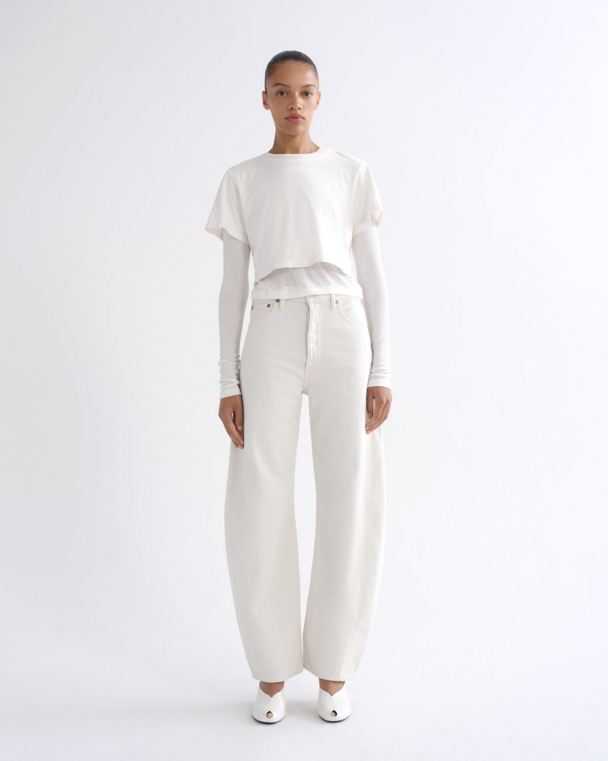A model stands against a seamless white studio background wearing a minimalist white outfit consisting of a long-sleeve cropped top, wide-leg trousers, and white pointed shoes.