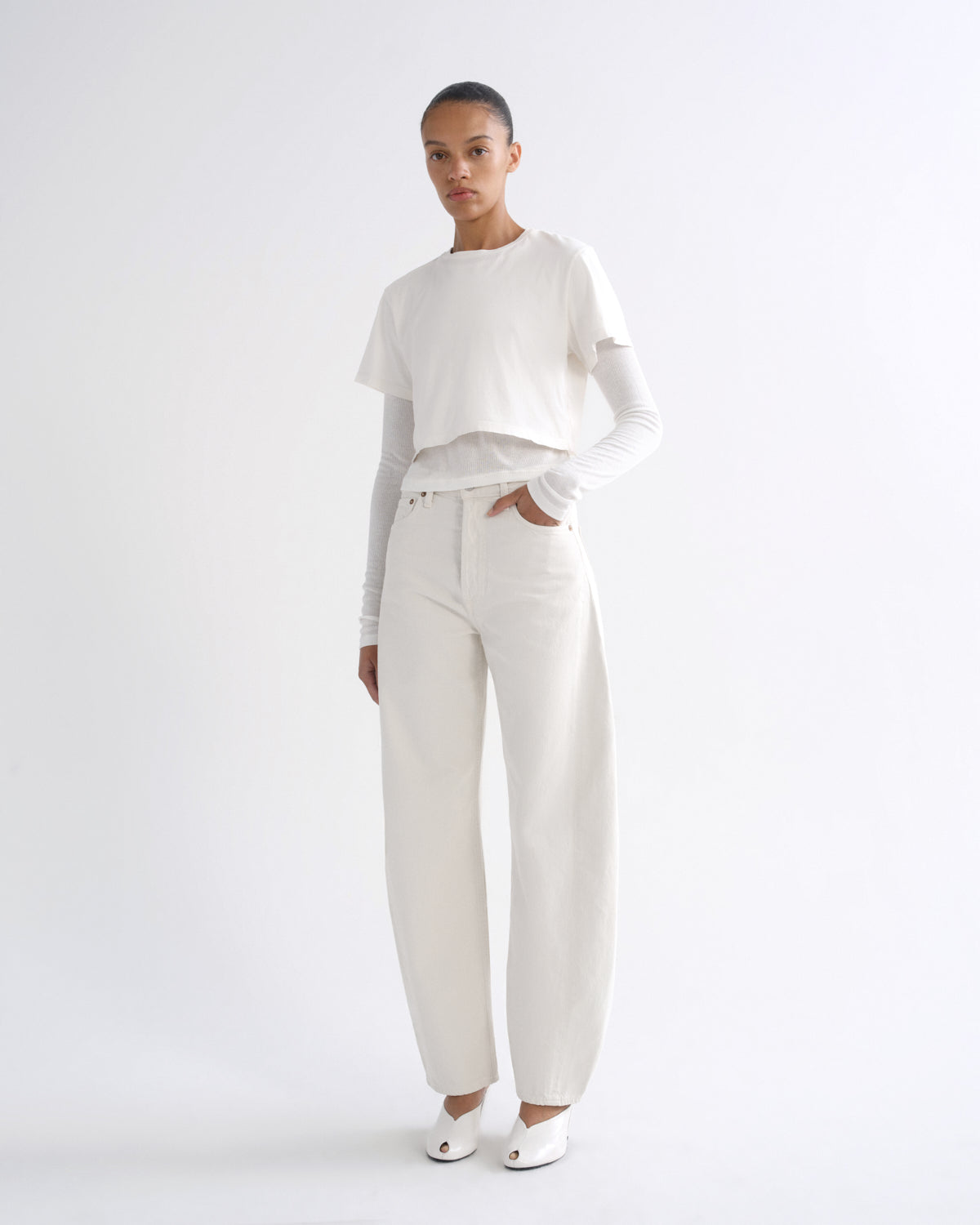 A model stands against a plain white studio background wearing a white short-sleeve cropped top over a fitted long-sleeve white shirt, white wide-leg pants, and white pointed shoes.