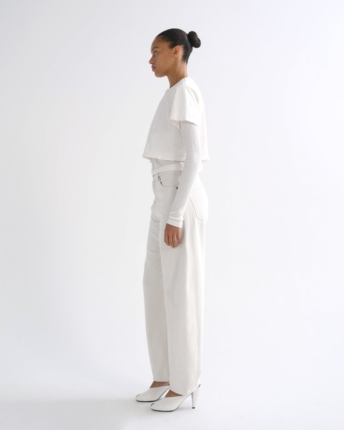 A model in profile wearing a white cropped long-sleeve top, white wide-leg trousers, and white pointed-toe heels against a plain white studio background.