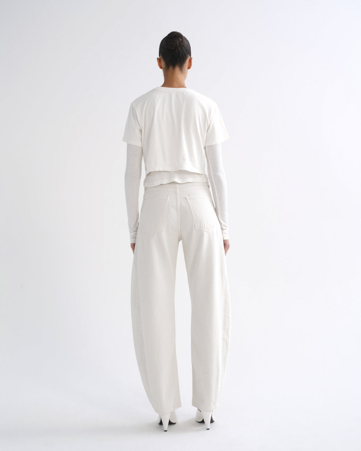 A model viewed from the back wearing a white cropped long-sleeve top over a white long-sleeve shirt, paired with wide-leg white trousers and white pointed shoes, against a plain white studio background.