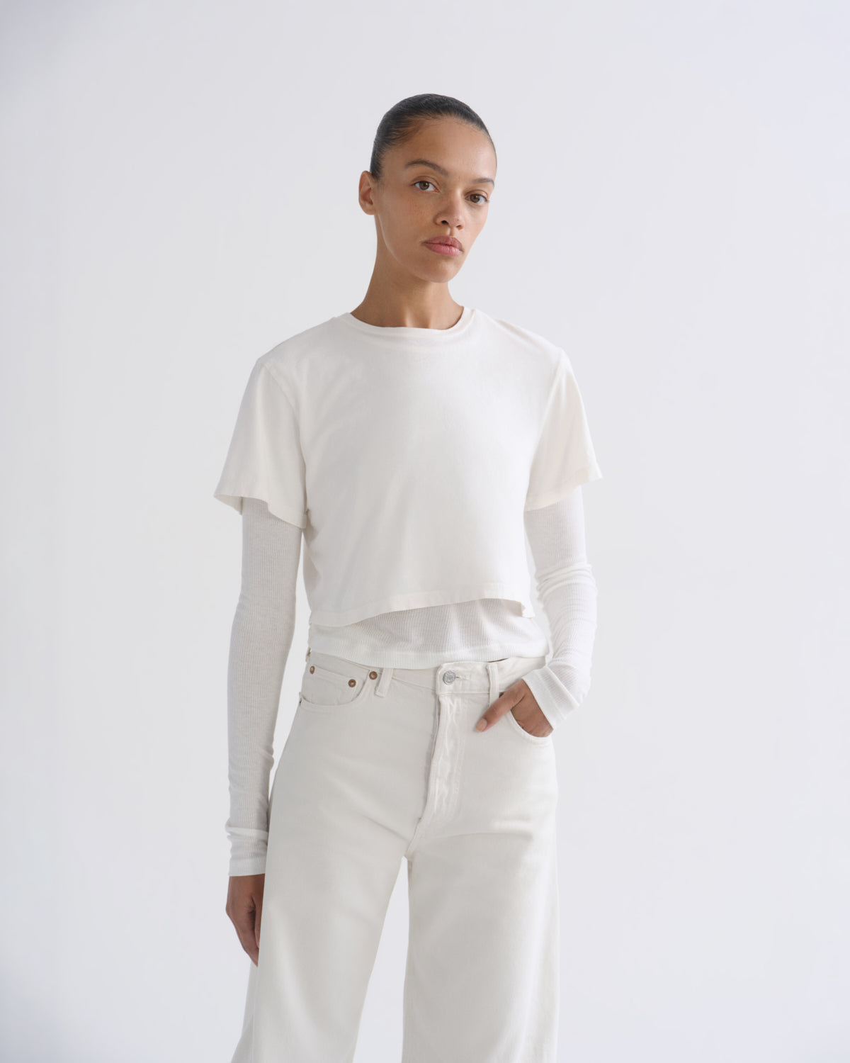 A model wearing a white short-sleeve cropped t-shirt layered over a white long-sleeve top, paired with white jeans, against a light gray studio background.